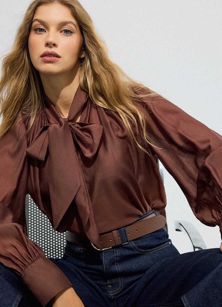 Femeie purtând o bluză elegantă cu fundă la gât, asortată cu blugi închiși la culoare și curea, prezentând o ținută smart casual stilată.