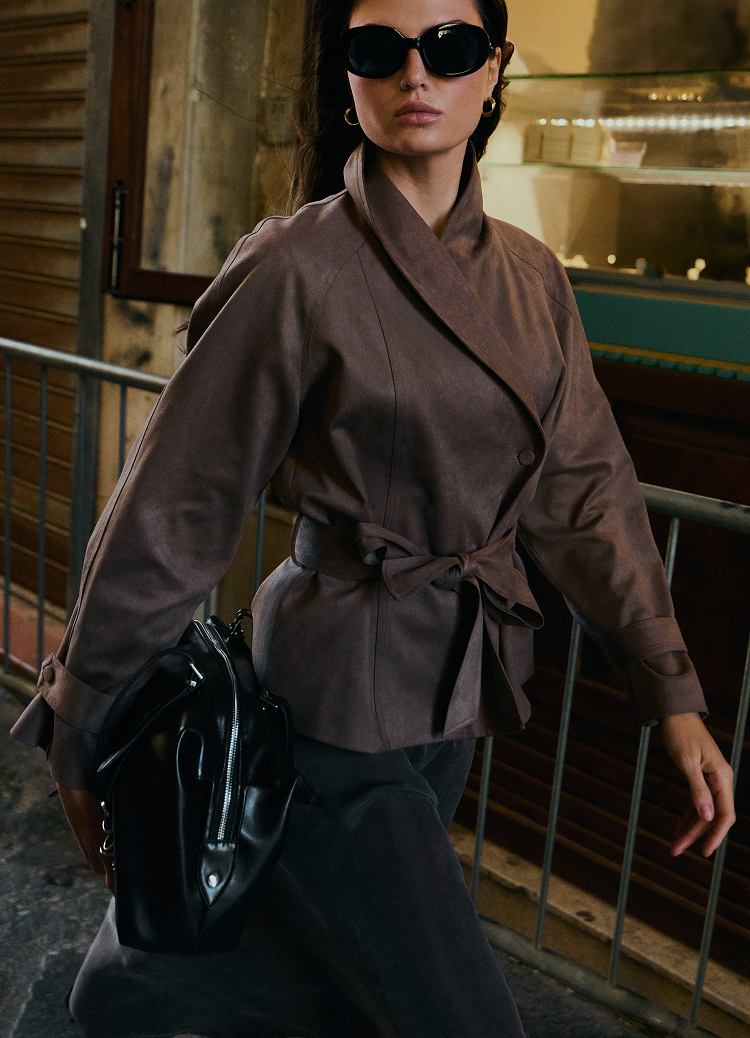 Promotivna modna slika koja prikazuje žensku jaknu sa pojasom, stilizovanu za svakodnevno urbano nošenje, uparenu sa aksesoarima koji prenose moderan i samouveren izgled.