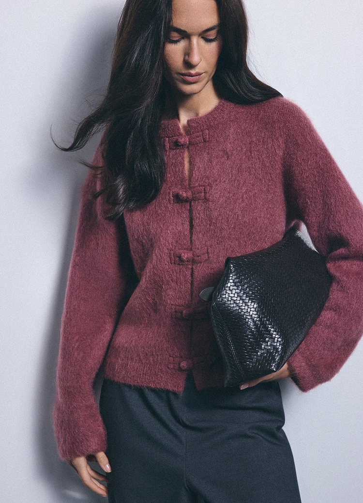 Žena nosi mekani pleteni kardigan s ukrasnim kopčama i drži pletenu clutch torbicu, predstavljajući elegantnu ležernu kombinaciju.