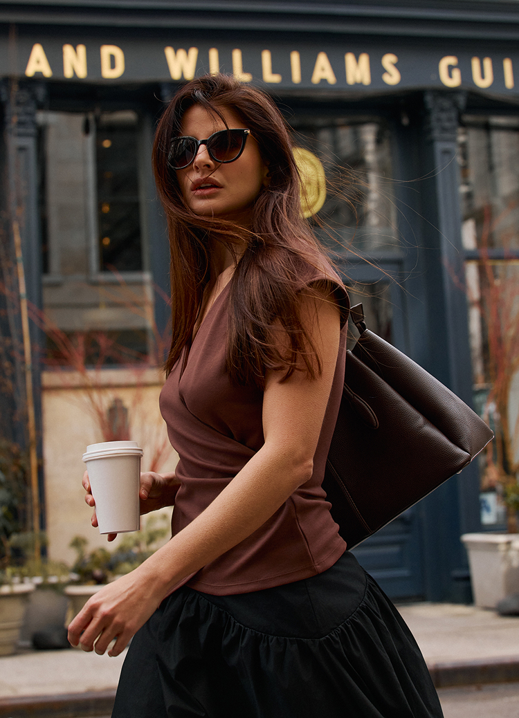 Osoba prolazi pored kafića, drži kafu za ponijeti i nosi veliku torbu, dočaravajući svakodnevni urbani stil života i trenutak u pokretu.